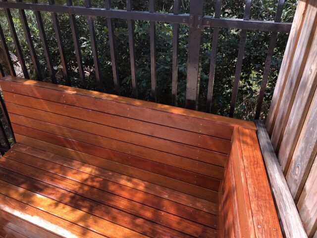 Stained hardwood built-in deck bench with backrest beside metal balustrade and greenery.