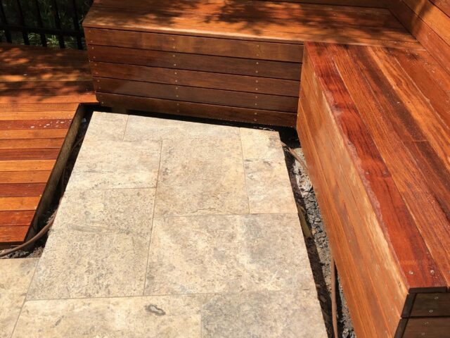 Hardwood built-in corner bench alongside travertine stone patio in sunlit backyard.