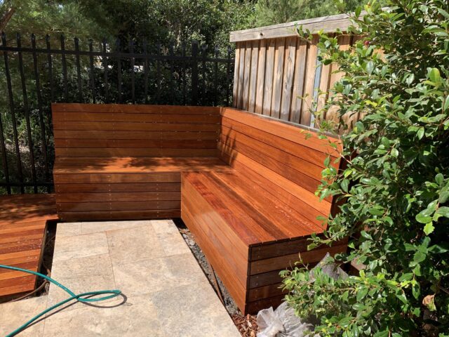 Built-in L-shaped hardwood bench on tiled patio beside shrubs and fence.