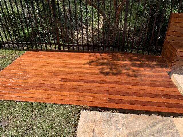 Low timber deck with built-in bench by black fence, beside lawn and stone pavers.
