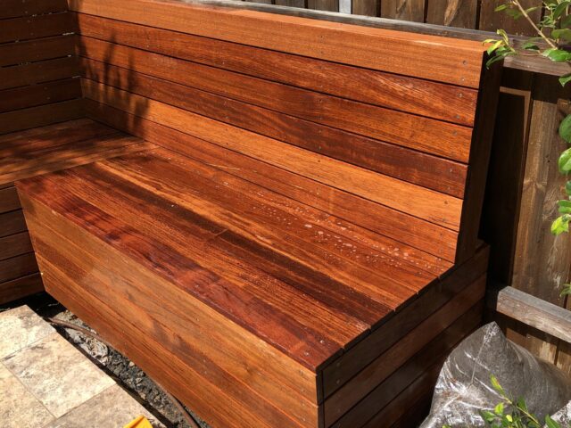Built-in hardwood L-shaped bench on tiled patio beside timber fence.