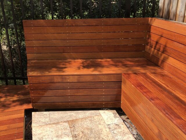 L-shaped built-in hardwood bench beside tiled patio and black metal fence.