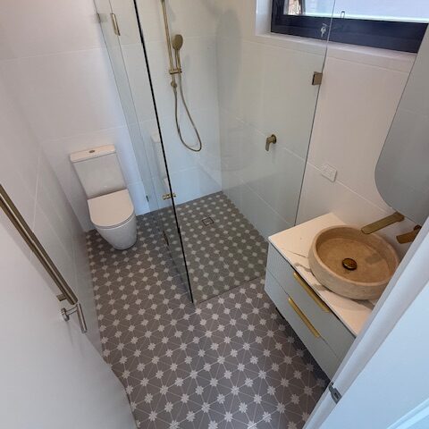 Compact bathroom with patterned grey tiles, frameless shower, brass fixtures, stone vessel sink, wall-hung vanity, toilet.