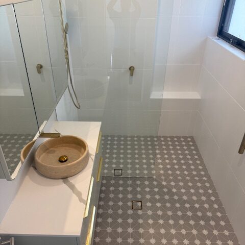 Renovated bathroom with patterned tile floor, frameless glass shower, brass fixtures, stone basin.