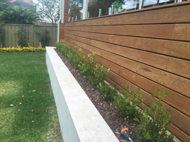 Horizontal timber-clad deck with glass posts, raised planter bed and lawn.