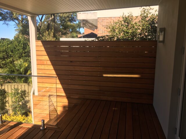 Undercover timber deck with glass balustrade, horizontal slat privacy screen, garden beyond.