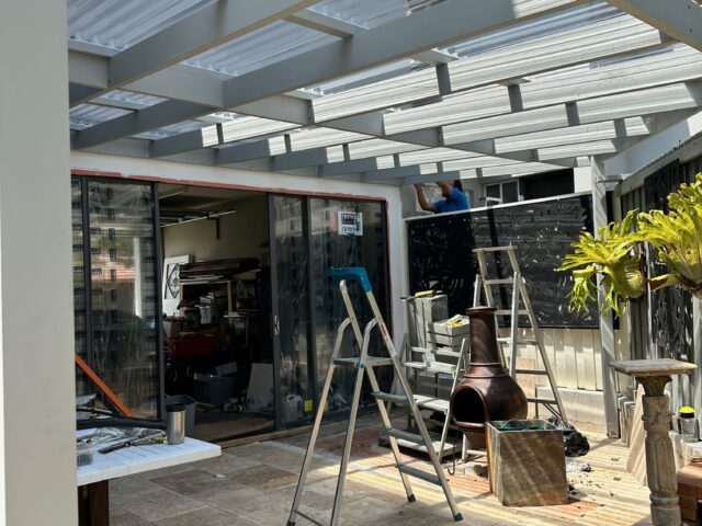 Builder installing clear polycarbonate pergola roof over tiled patio, ladders and tools.