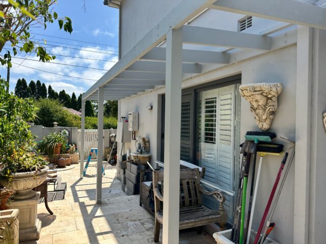 Light grey pergola frame under construction over tiled patio with ladder, tools and plants.