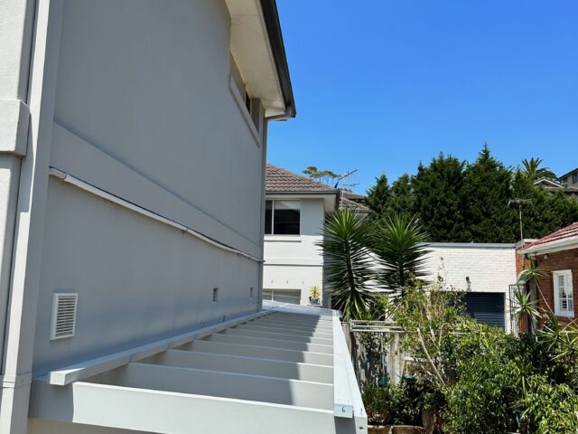 Pergola frame attached to side of home above paved courtyard and garden.