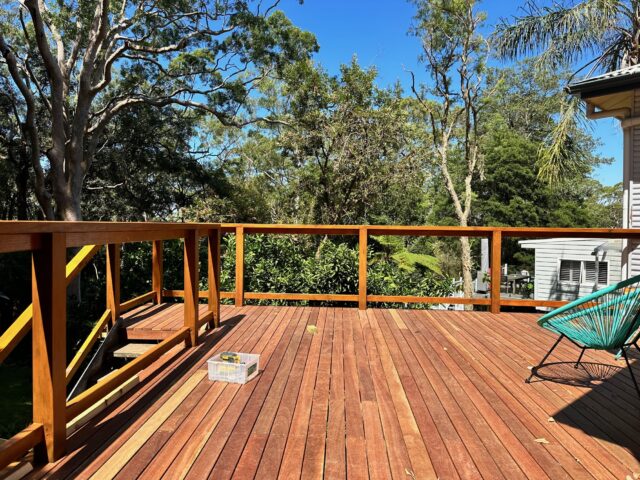 Sunlit hardwood deck with timber railing, steps, turquoise chair, and leafy garden outlook.
