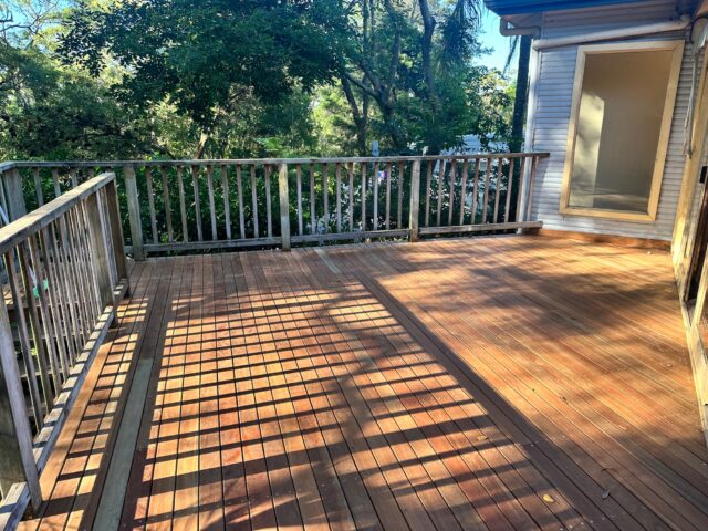 Spacious sunlit timber deck with railings beside house, overlooking leafy bushland.