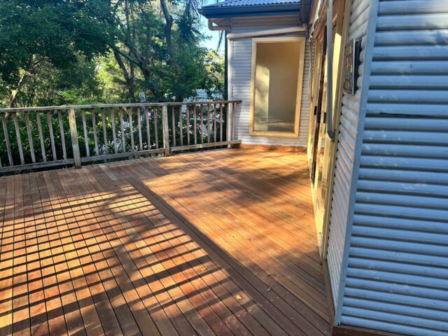 Sunlit timber deck with balustrade beside corrugated-clad house, surrounded by trees.