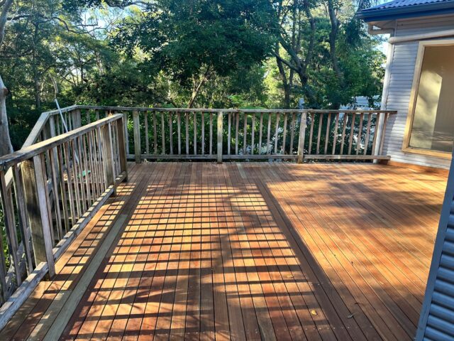 Elevated timber deck with railing beside house, dappled sunlight, trees surrounding.