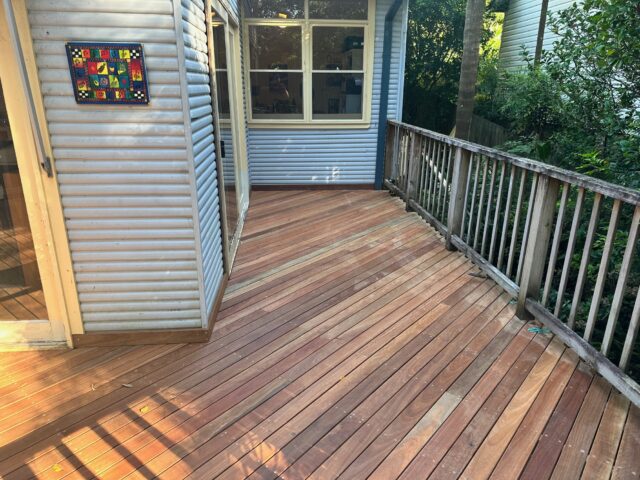 Corner timber deck with angled boards, adjacent weatherboard house and railing facing leafy backyard.