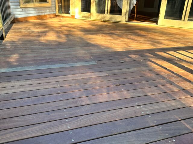 Hardwood timber deck adjoining house with sliding glass doors, sunlit with railing shadows.
