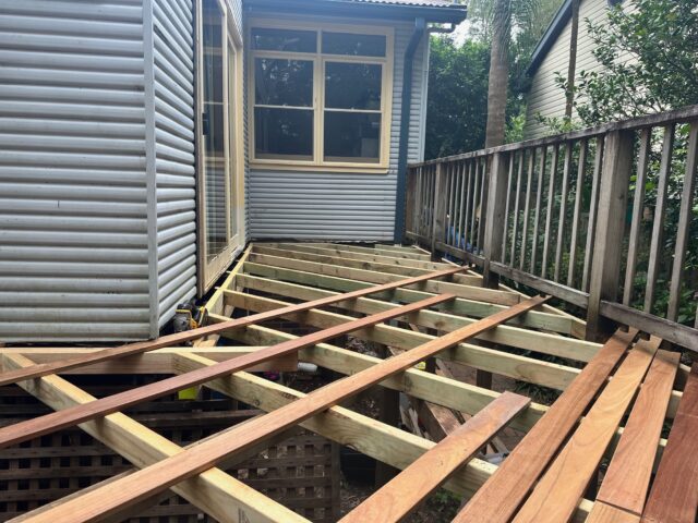 Timber deck framing with hardwood boards being installed beside corrugated-clad house and balustrade.