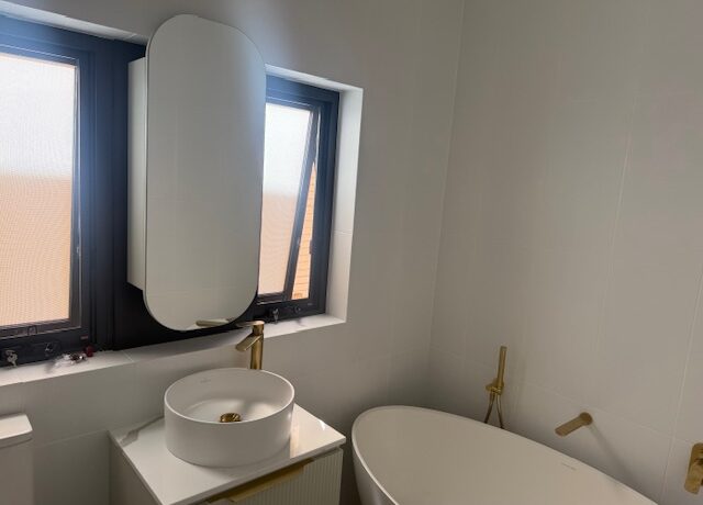 White-tiled bathroom with freestanding tub, round vessel sink, gold fixtures and black-framed windows.