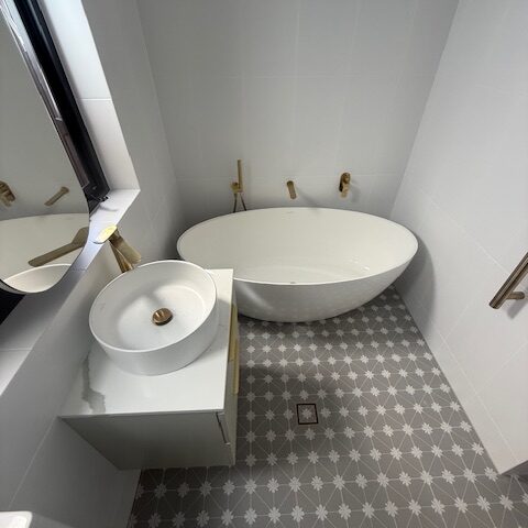Freestanding oval bathtub, vessel sink vanity, brass tapware, grey star-patterned tiles, central floor drain.