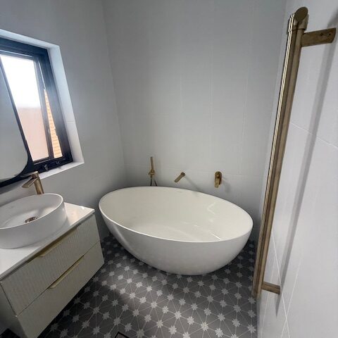 Renovated bathroom with freestanding tub, wall-mounted vanity, brass fixtures, patterned grey floor tiles.