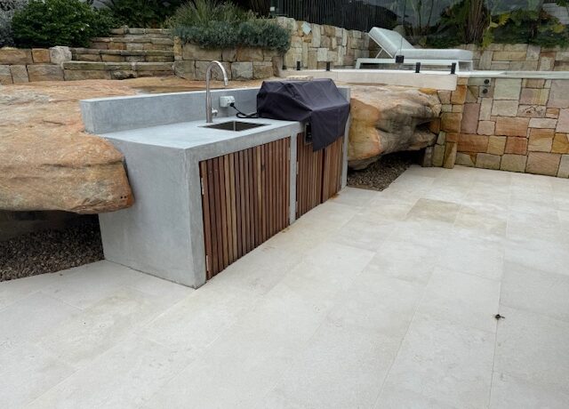 Outdoor kitchen with concrete benchtop, sink and covered BBQ set into sandstone.