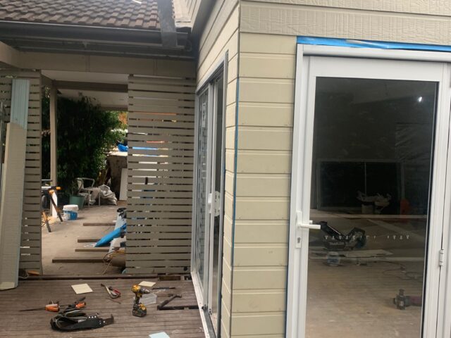Timber deck under construction beside new clad extension with glass doors, slatted screen, and tools on decking.