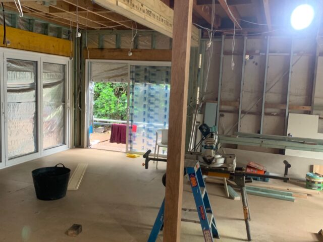 Open-plan home renovation with exposed timber framing, hanging wiring, sliding doors, ladder and mitre saw.