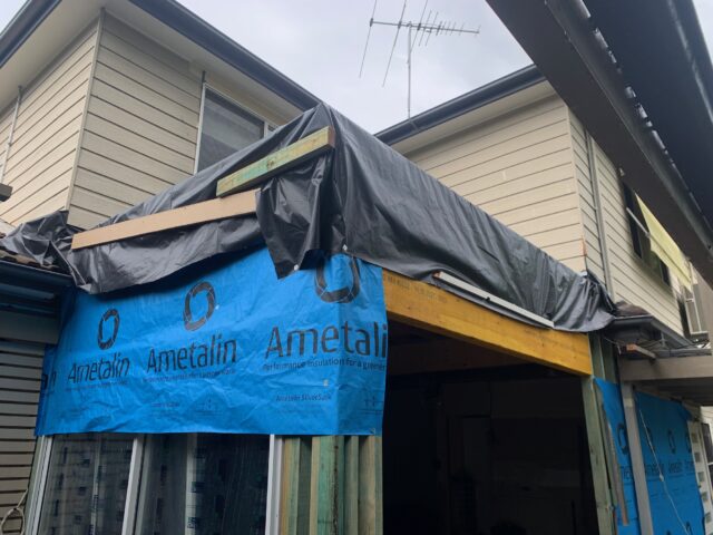 Timber-framed home extension with blue building wrap and temporary tarp roofing.