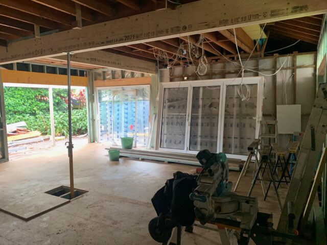 Home renovation in progress with exposed framing, sliding glass doors to garden, hanging wiring, and miter saw.