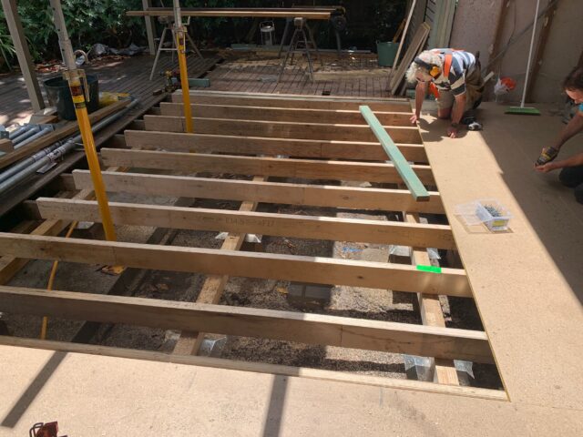 Two builders installing flooring sheets over timber joists for an outdoor deck.