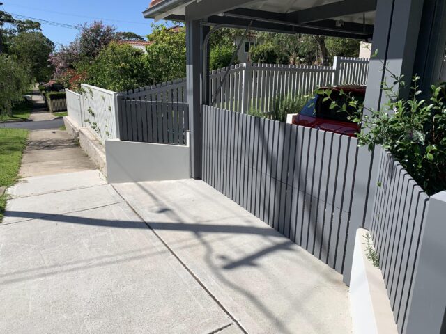 Stepped grey slat boundary fence with carport beside sloped concrete driveway.