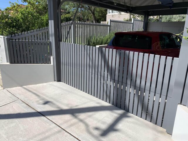 Modern grey vertical slat driveway gate and fence, carport sheltering red car