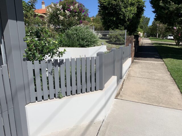 Grey picket fence on rendered retaining wall bordering a landscaped front garden and footpath.
