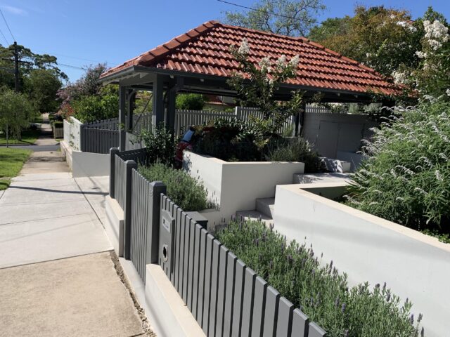 Landscaped front yard with grey picket fence, rendered retaining walls and tiled-roof carport.