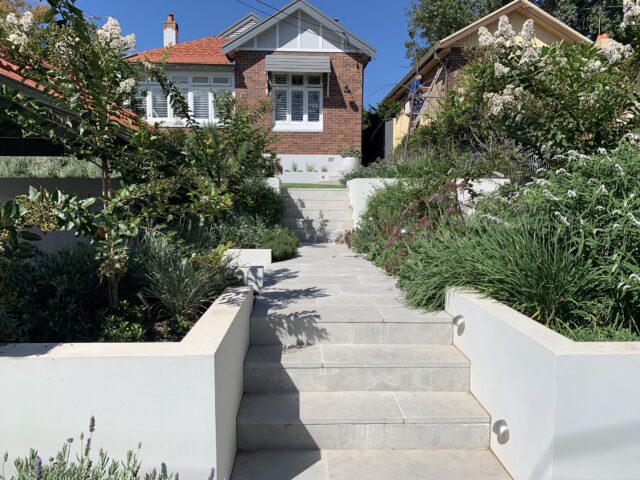 Rendered retaining walls, stone steps and path lighting through landscaped front garden to brick bungalow.