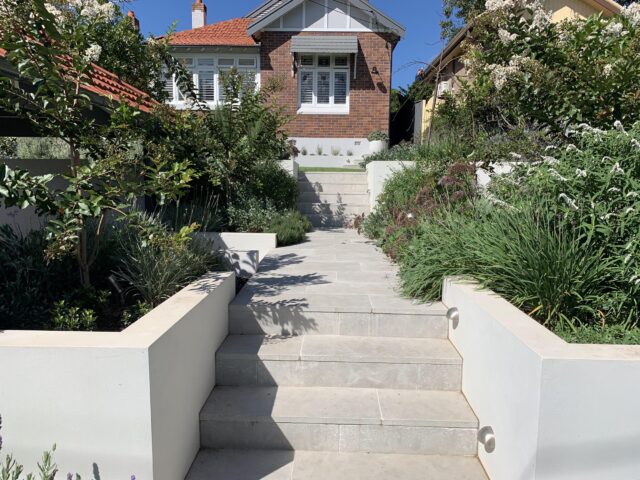 Terraced stone steps with rendered retaining walls, garden lighting and lush planting to brick home.