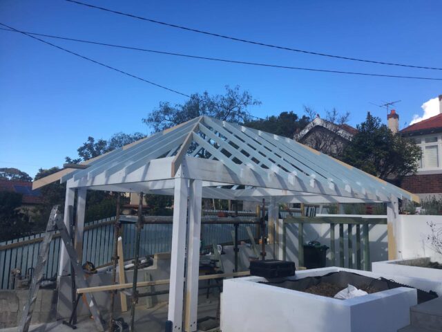 Backyard gable pergola roof framing in progress with white posts, rafters and scaffolding.