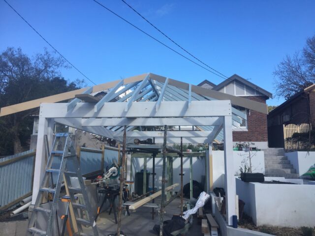 Timber pergola frame under construction, pitched rafters, ladder and tools in suburban backyard.