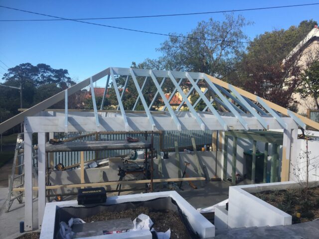 Timber gable roof framing underway over concrete slab in residential yard, tools and ladders visible.