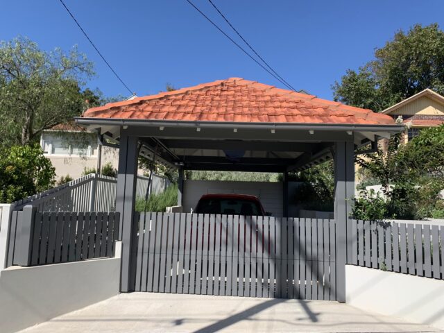 Terracotta-tiled carport with grey slatted fence and gate, red car parked.