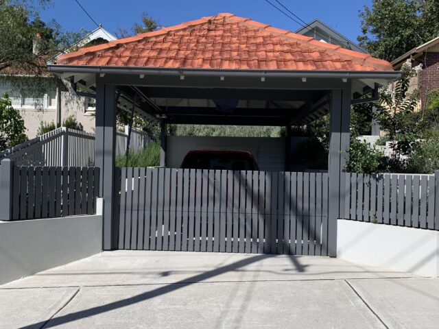 Single-car carport with terracotta tiled hip roof, grey slatted gate and concrete driveway.