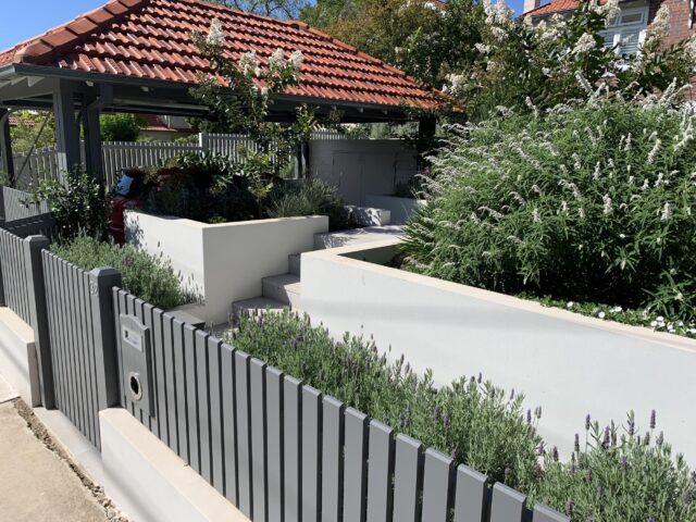 Tiered rendered planters, steps and lush landscaping beside grey slat fence and tiled-roof carport.