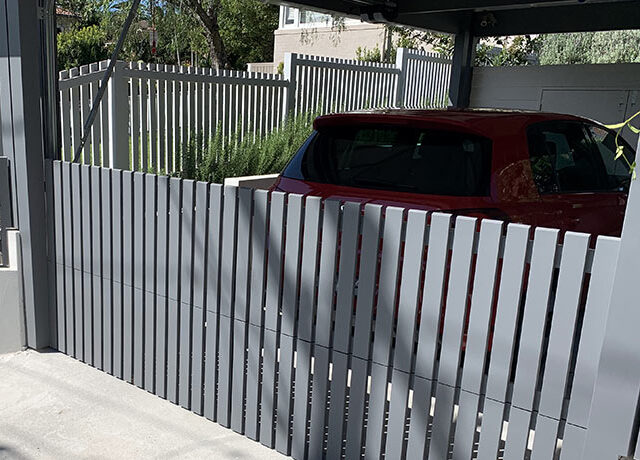 Modern grey aluminium slat fence and gate enclosing carport with red car.
