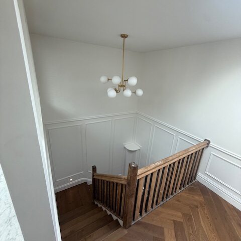 Staircase with herringbone timber floor, white wainscoting, and brass globe chandelier.