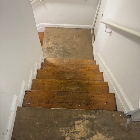 Narrow indoor timber stairs with white handrails and a scuffed subfloor landing.