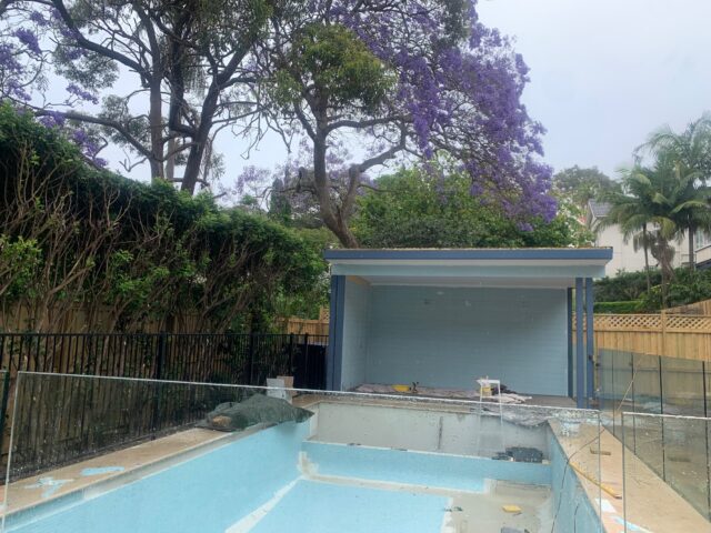 Backyard pool renovation with cabana construction, glass fence, hedges and flowering jacaranda.