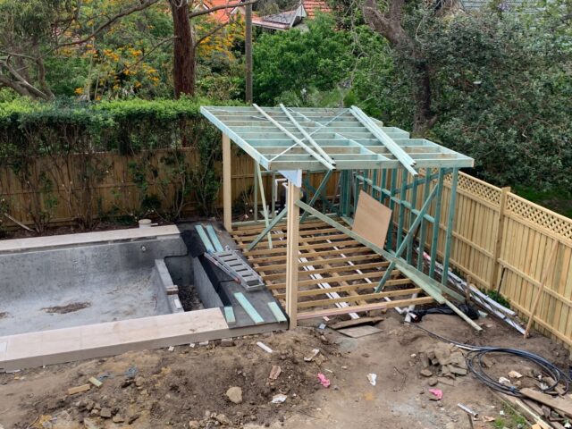 Timber deck and roof framing beside an empty concrete pool, backyard renovation.