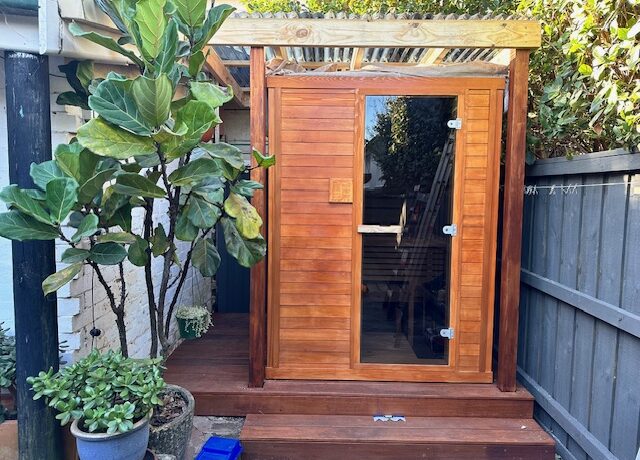 Compact timber-clad garden room with glass door and polycarbonate pergola, on a small deck with potted plants.