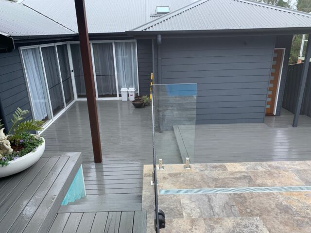 Grey composite deck with planter, glass pool fence, travertine tiles, and sliding doors.