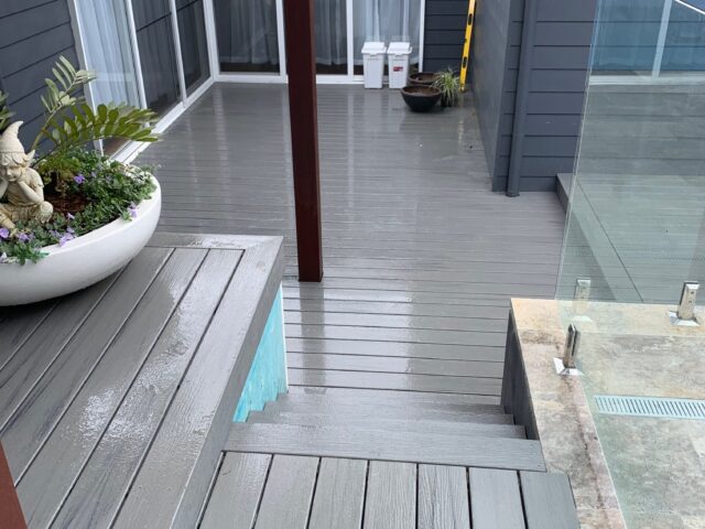 Wet grey decking with steps, planter, and glass pool fence by sliding doors.