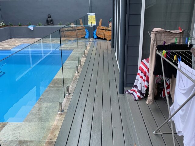 Narrow grey composite deck along house beside pool with frameless glass fence, landscaped retaining wall.